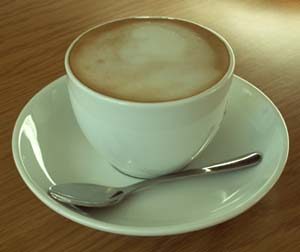 A white cup of coffee with froth--crema--sits atop a white saucer; a small silver spoon rests in the saucer
