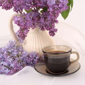 White vase filled with lilacs sits behind a brown cup of coffee, sitting in a saucer