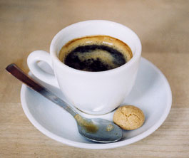 Awhite cup of dark coffer with a ring of crema; in the saucer sit a small spoon and a tea cookie