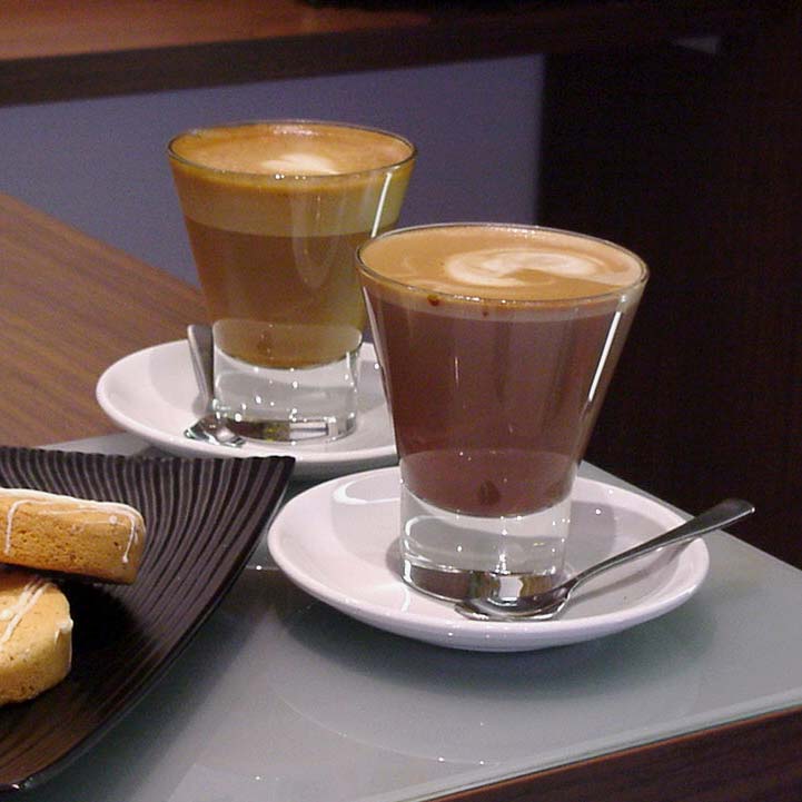 A table with grey placemat holds two white saucers, each containing a glass filled with coffee. In the front saucer rests a small, silver spoon. To the left, dark brown plate with biscotti rests.