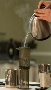 Three metal cups sit on a white stove; a kettle pours steaming water through a filter atop the center cup
