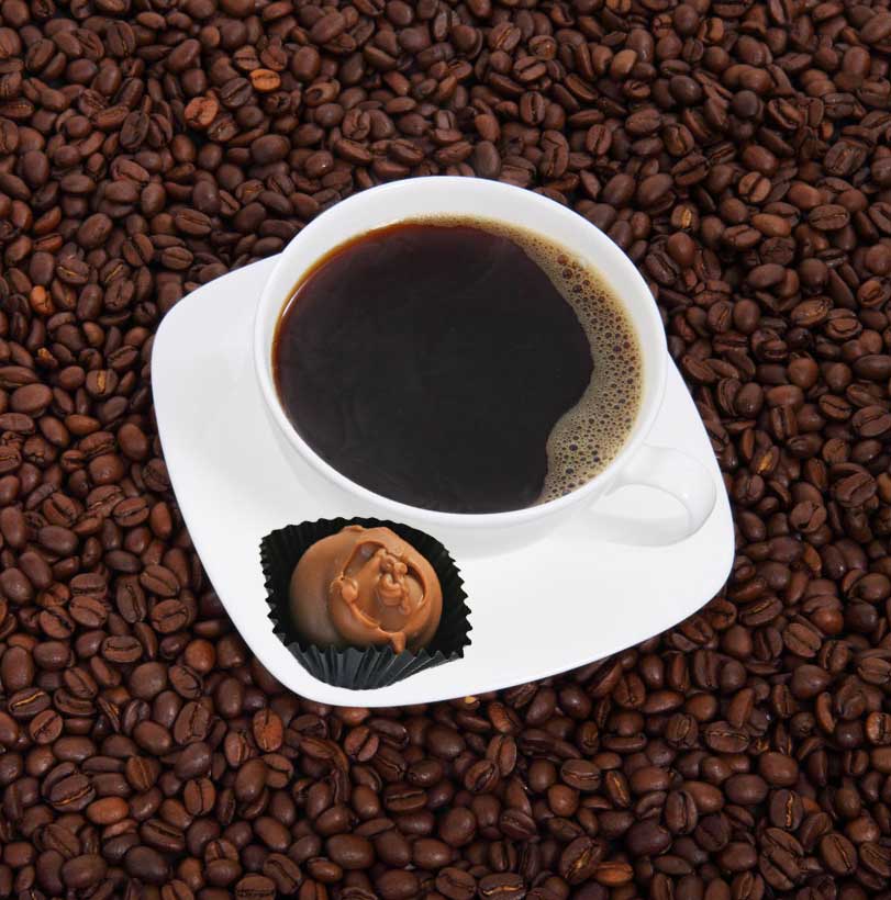 A white cup, filled with black coffee, sits in a matching saucer atopa field of roasted coffee beans; in the saucer rests a chocolate truffle candy.