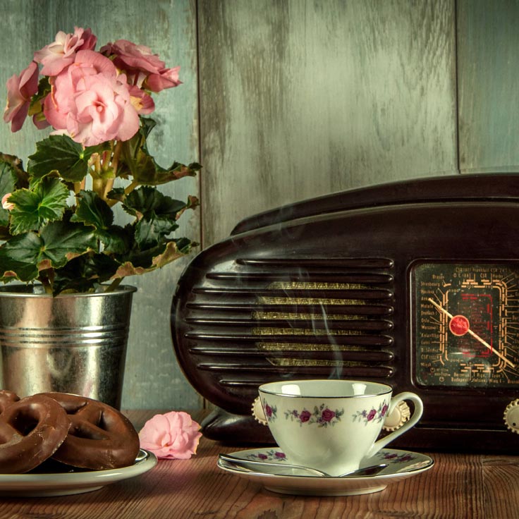 On a wooden table sit apotted miniature pink rose, a vintage radio and a china coffee cup.